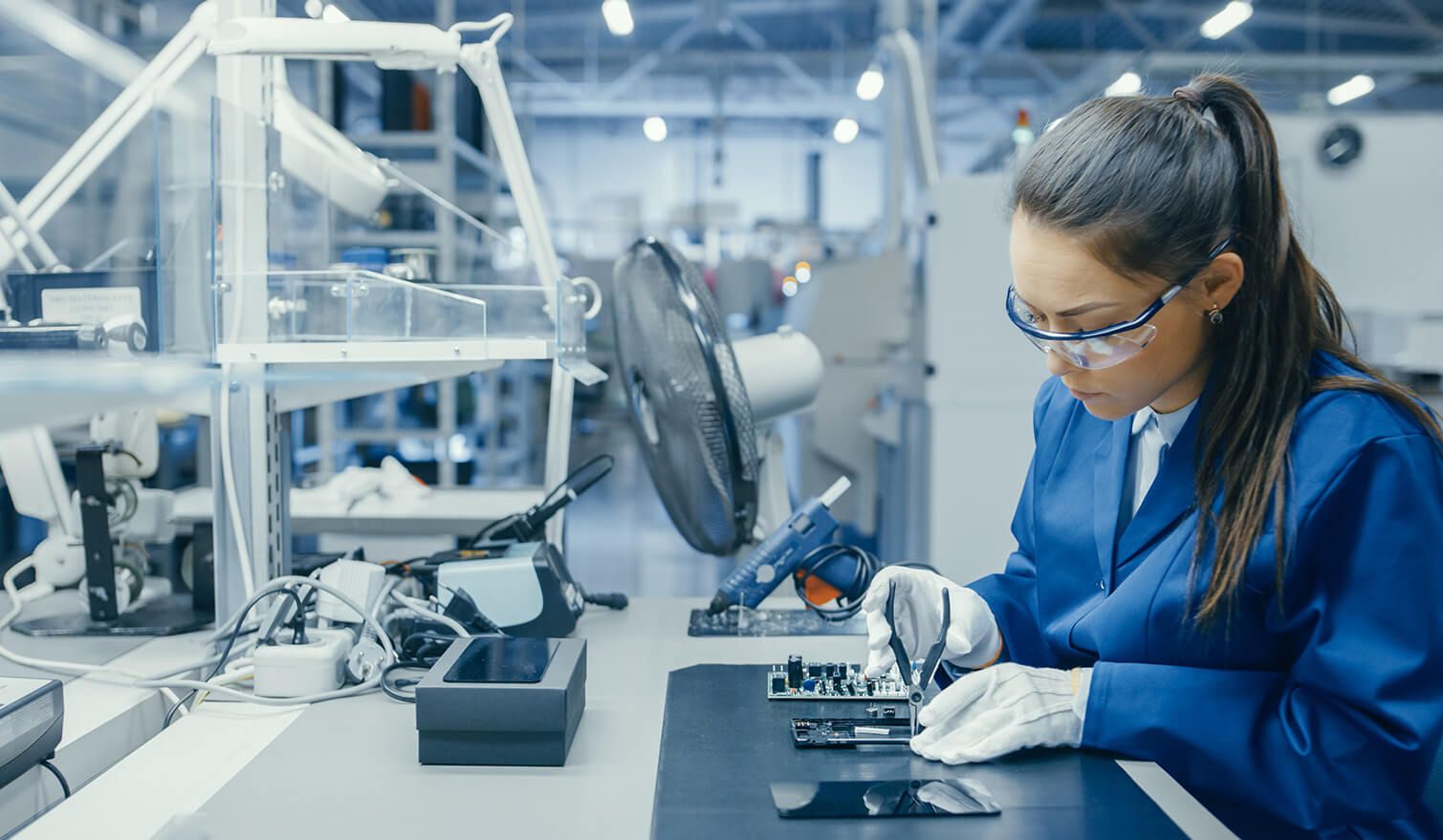 Eine Frau in blauer Arbeitskleidung bedient präzise elektronische Geräte in einer entsprechend beleuchteten Produktionsumgebung
