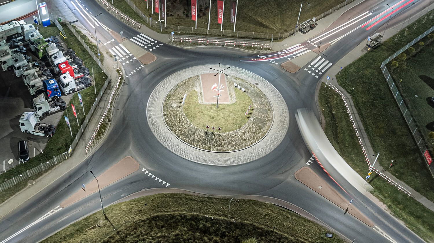 Nachtansicht aus der Vogelperspektive auf einen von Straßenlaternen beleuchteten Kreisverkehr, die den Autoverkehr sowie die Beschilderung zur Kennzeichnung der Fahrtrichtung zeigt.